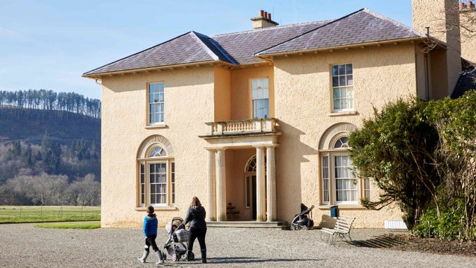 A child and adult, pushing a pram, walk past the entrance of the villa at Llanerchaeron, Wales